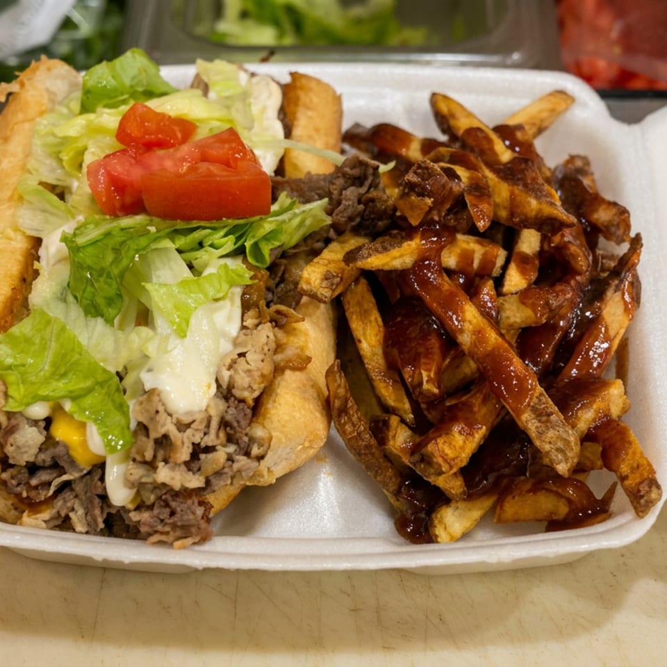 Cheesesteak sandwich topped with lettuce, tomato, and mayonnaise beside barbecue-sauced fries in a foam takeout container.