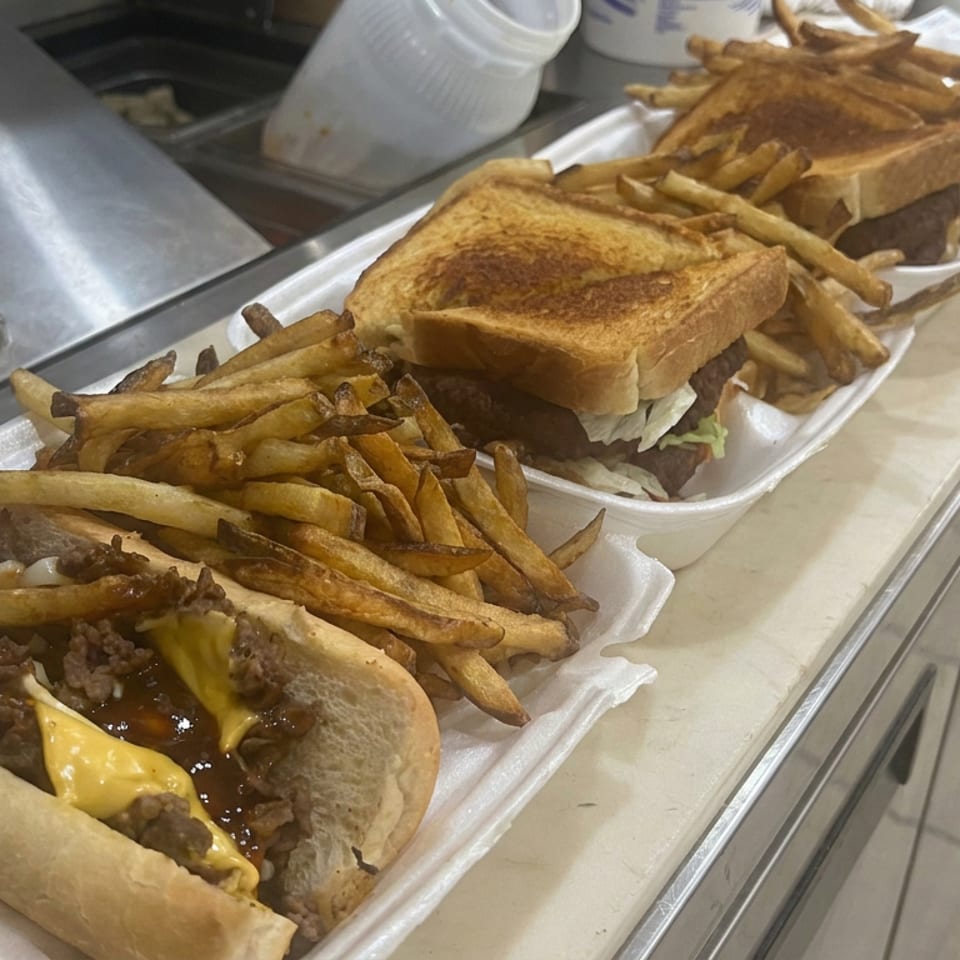 Three takeout trays hold toasted beef sandwiches, including a cheesesteak with melted cheese, alongside piles of French fries.